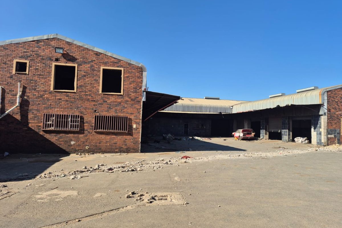 Rear view of a large industrial-style brick building featuring metal roofing, security bars, and an expansive concrete yard area requiring significant maintenance.