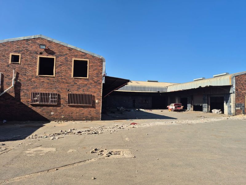 Rear view of a large industrial-style brick building featuring metal roofing, security bars, and an expansive concrete yard area requiring significant maintenance.