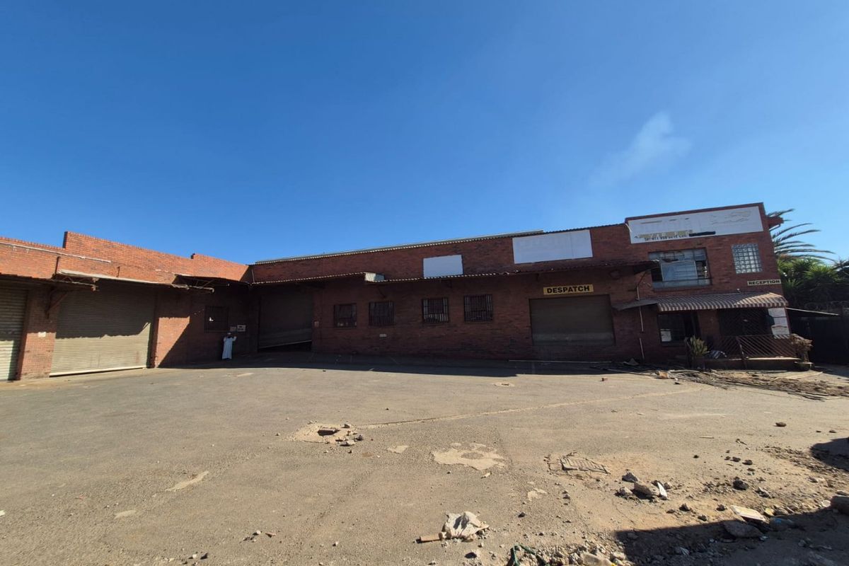 Industrial property exterior featuring a large asphalt yard, multiple roller shutter doors, and brick facade construction with visible maintenance requirements.