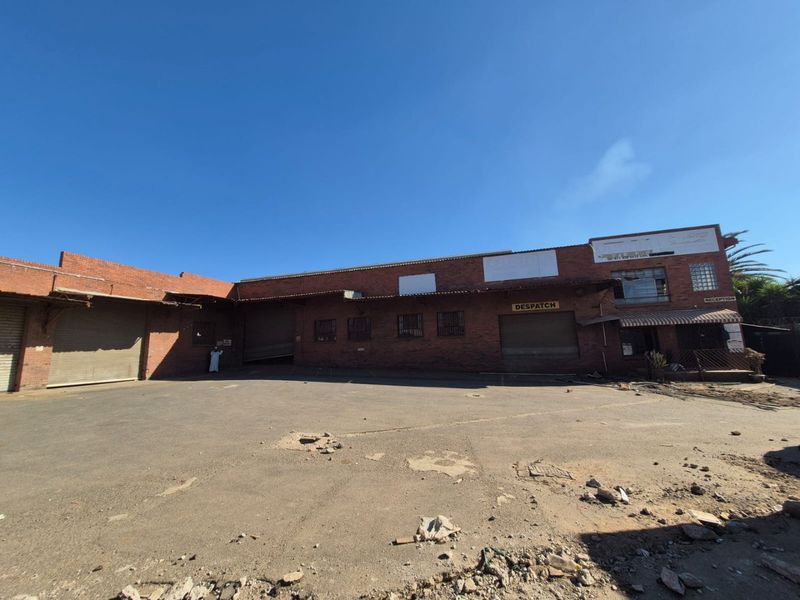 Industrial property exterior featuring a large asphalt yard, multiple roller shutter doors, and brick facade construction with visible maintenance requirements.
