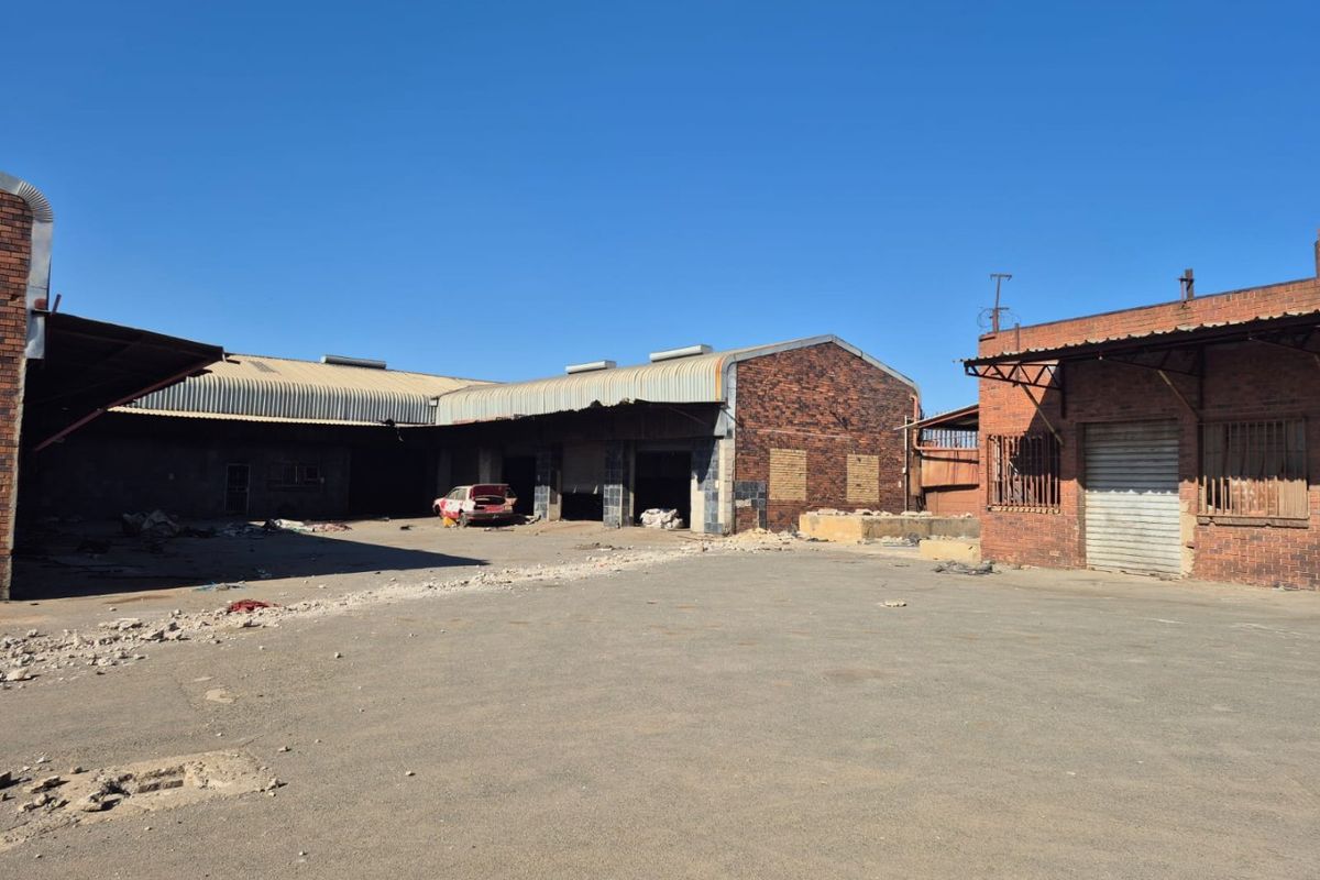 A large industrial-style rear yard featuring brick buildings with corrugated metal roofing, roller shutter doors, and significant debris requiring site clearance.