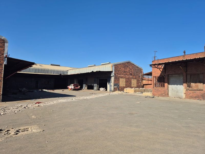 A large industrial-style rear yard featuring brick buildings with corrugated metal roofing, roller shutter doors, and significant debris requiring site clearance.