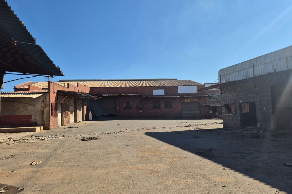 An industrial exterior yard area featuring brick warehouse buildings with corrugated metal roofing and roller shutter doors, showing significant signs of wear and deterioration.