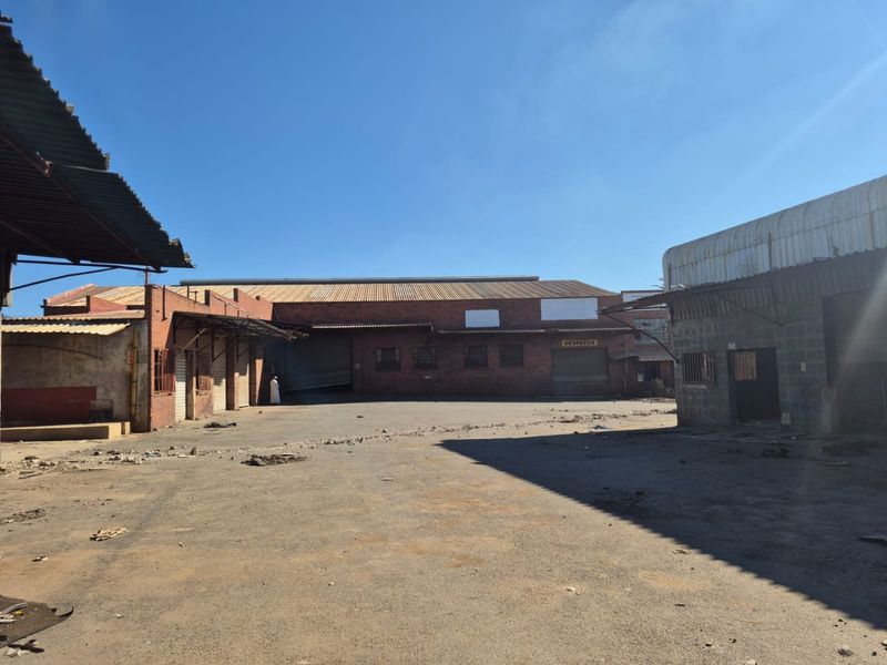 An industrial exterior yard area featuring brick warehouse buildings with corrugated metal roofing and roller shutter doors, showing significant signs of wear and deterioration.