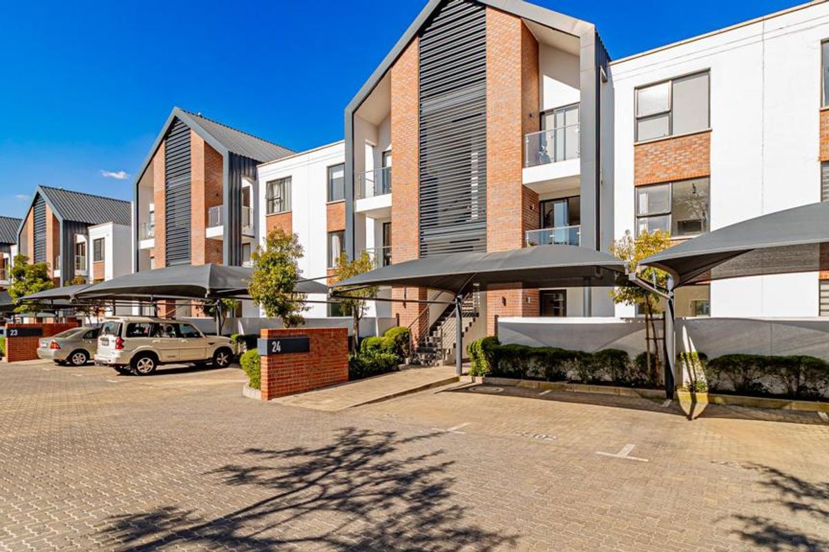 Modern apartment complex exterior featuring brick and white facade, paved parking area with shade cloth carports, and private balconies with glass balustrades.