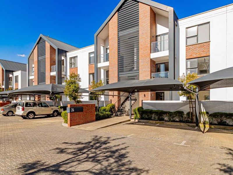Modern apartment complex exterior featuring brick and white facade, paved parking area with shade cloth carports, and private balconies with glass balustrades.