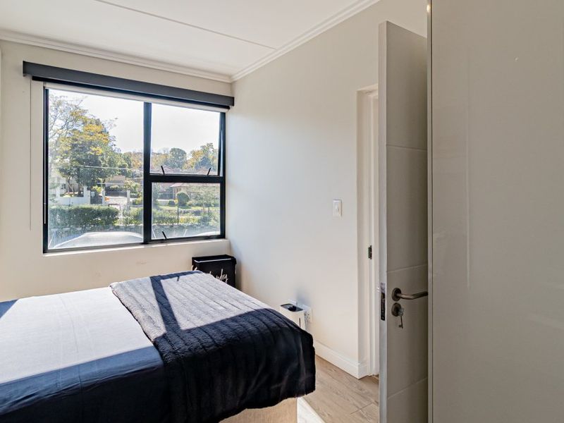A bright bedroom featuring laminate flooring, a built-in wardrobe, and large windows providing natural light and a pleasant outdoor view.