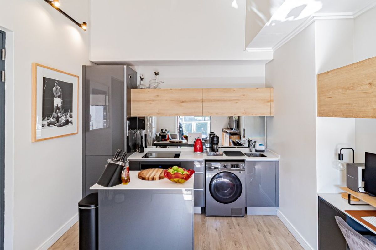 Modern compact kitchen featuring grey cabinetry, wood-look upper cabinets, integrated washing machine, mirror splashback, and an adjacent built-in workspace.