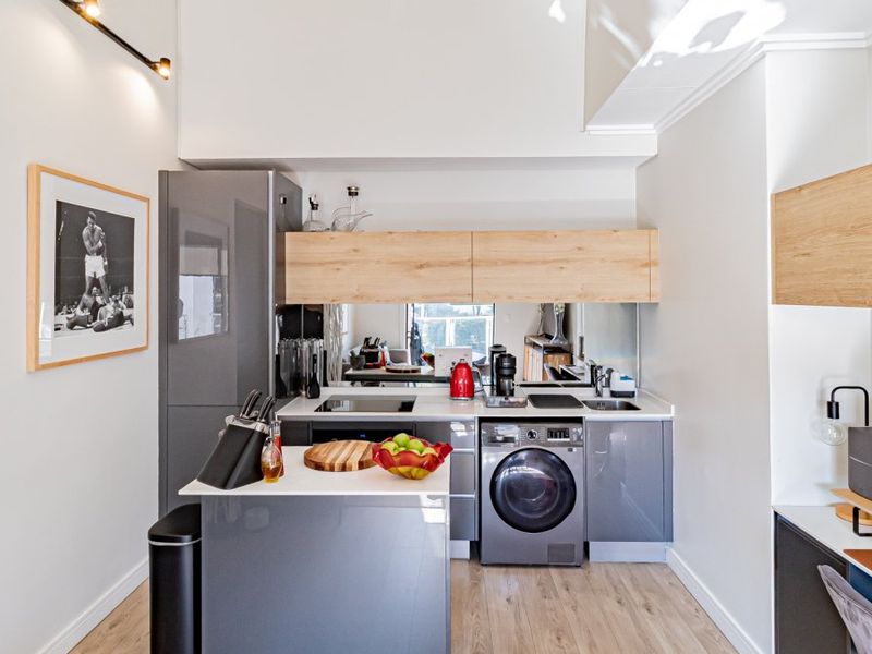 Modern compact kitchen featuring grey cabinetry, wood-look upper cabinets, integrated washing machine, mirror splashback, and an adjacent built-in workspace.
