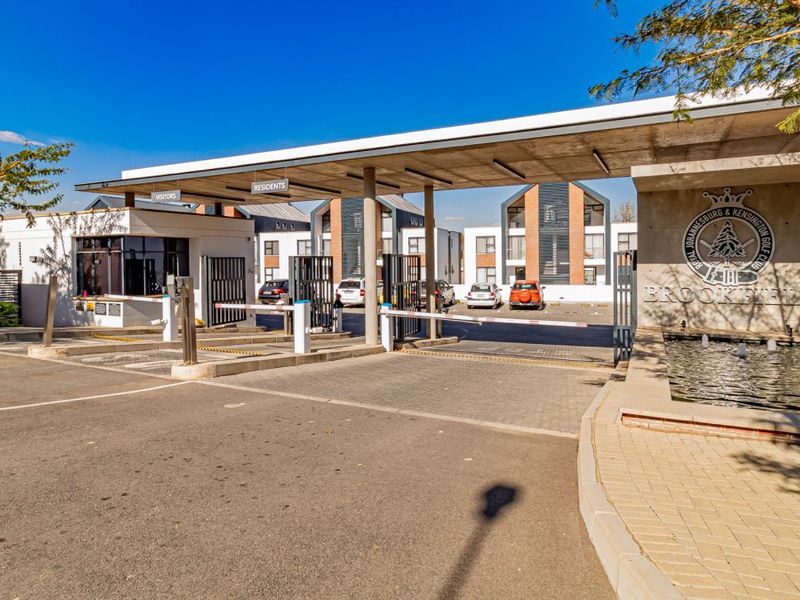 The secure entrance gatehouse of the Brookfield apartment complex, featuring a modern canopy, boom gates, and a decorative water feature.