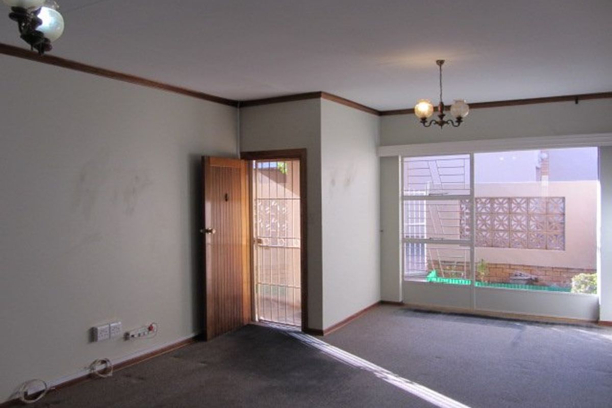 An empty living room featuring carpet flooring, a timber door with security gate, and large windows providing natural light.