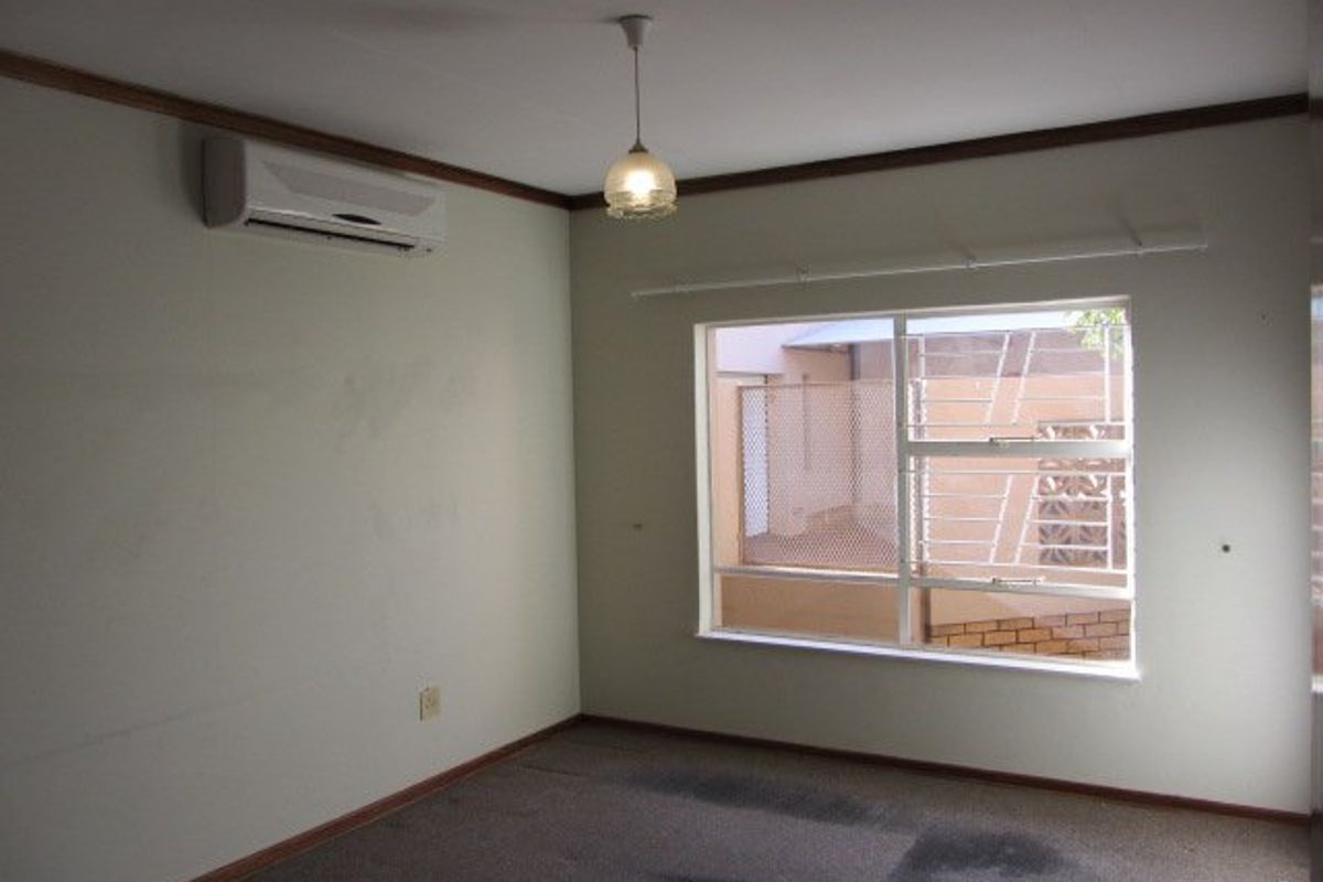 An empty bedroom featuring a split-system air conditioner, pendant lighting, and a window with security bars. The room requires cosmetic updates and carpet cleaning.