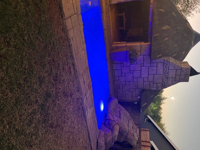 Outdoor entertainment area featuring a swimming pool with a rock feature, adjacent to a thatched roof lapa with stone cladding.