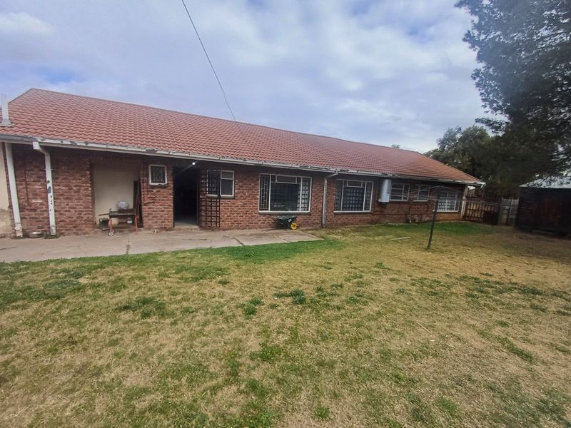 Rear view of a single-story brick house featuring a tiled roof, concrete patio area, and established garden space.