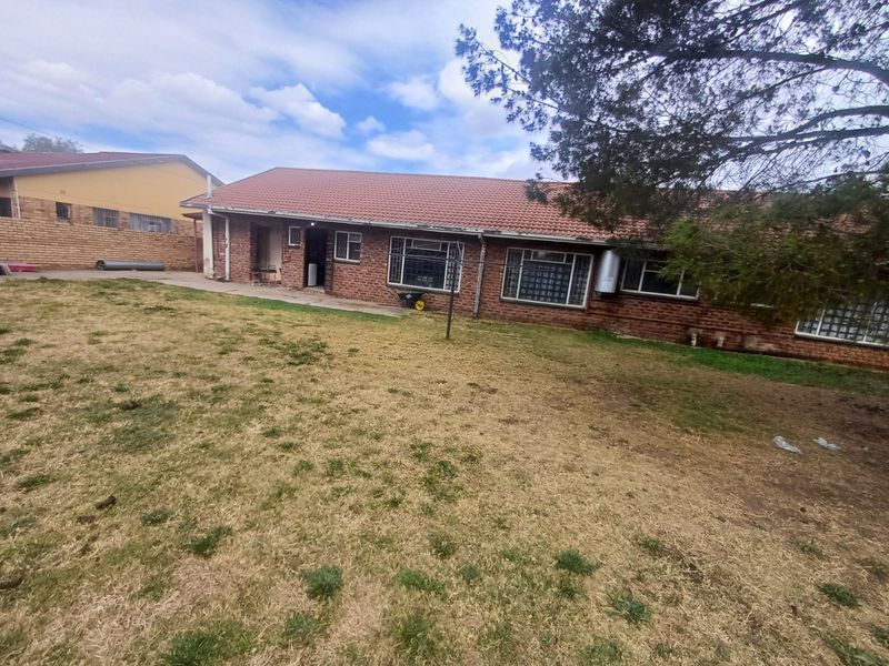 Rear view of a single-story brick house featuring a tiled roof, security bars on windows, and an external geyser, set against a large lawn.