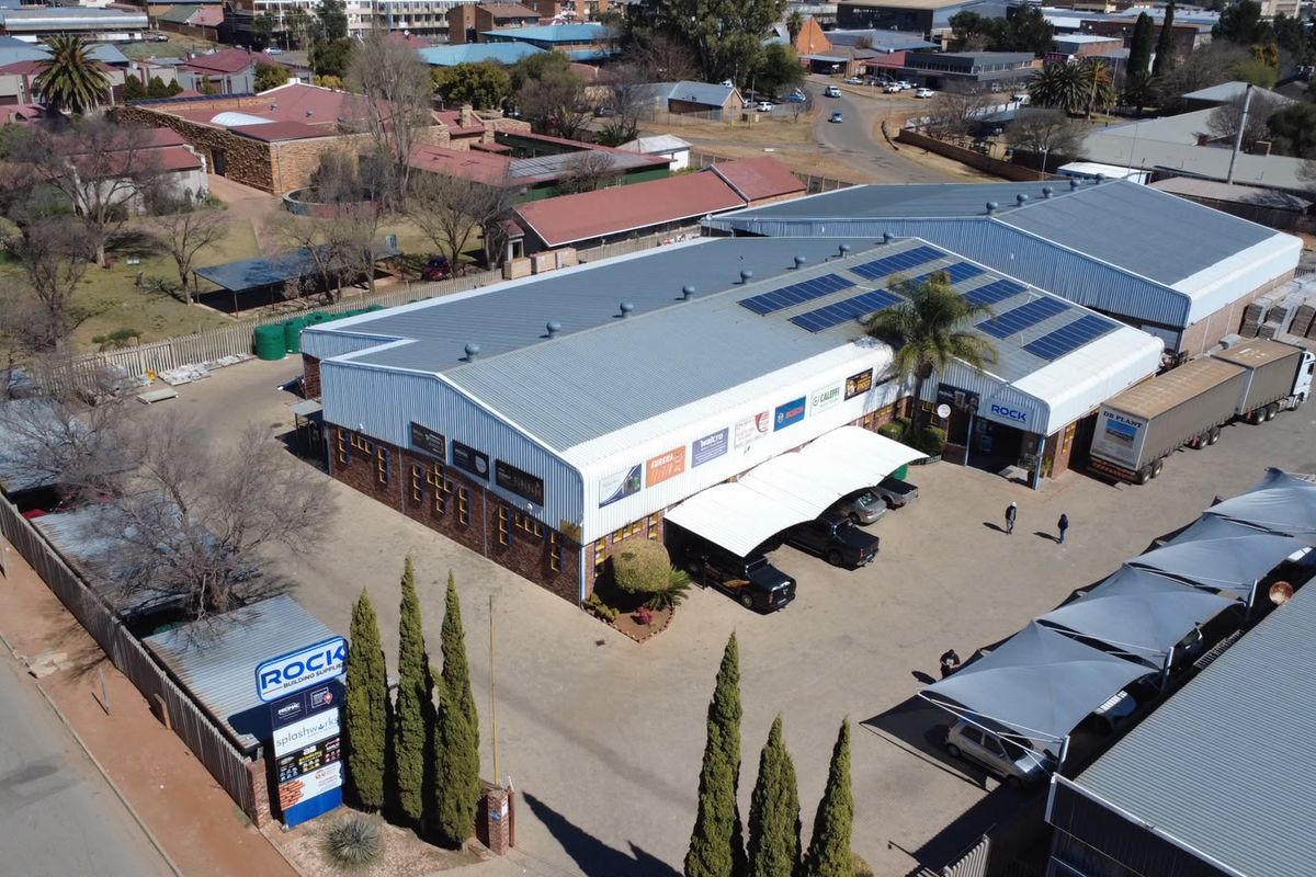 Aerial view of a commercial building complex featuring metal roofing, solar panel installations, paved parking areas with shade structures, and perimeter fencing.