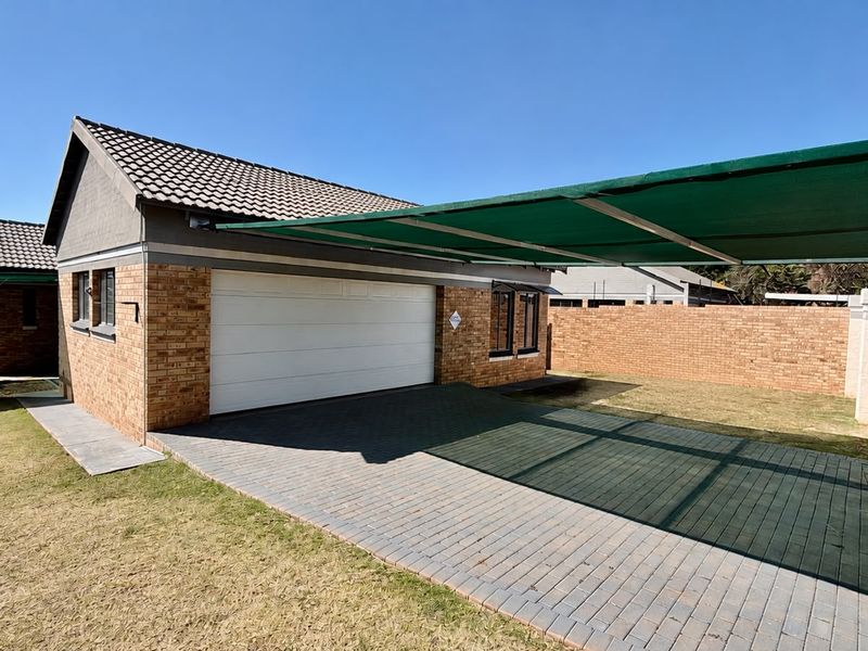 Front view of a townhouse featuring a double garage, paved driveway, and a shade cloth carport, set within a neat, walled property boundary.