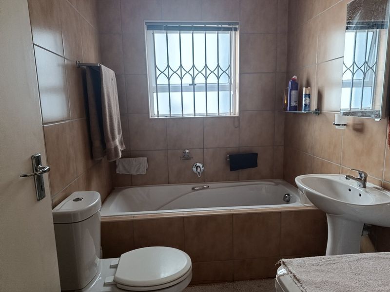A functional bathroom featuring a bathtub, pedestal basin, and toilet, finished with neutral wall tiles and security bars on the window.