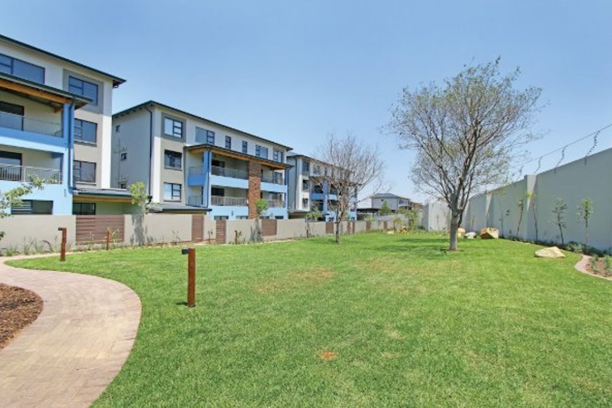 A well-maintained communal garden area featuring a paved walkway, manicured lawn, and perimeter wall with electric fencing, adjacent to the apartment complex.