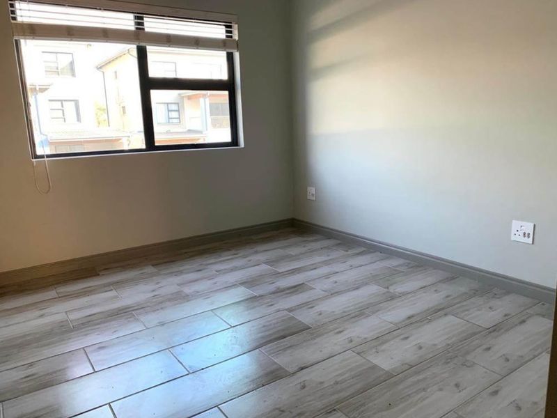 A bright, empty bedroom featuring modern wood-look tiled flooring, neutral walls, and a large window with fitted blinds.