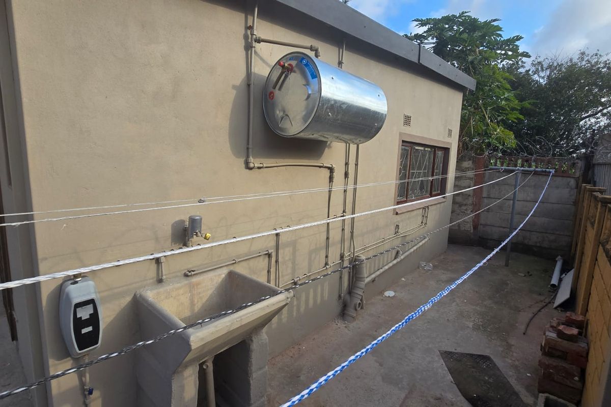 Rear exterior utility area featuring a wall-mounted electric geyser, concrete wash trough, and exposed plumbing infrastructure against a rendered wall.