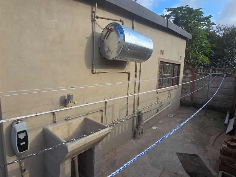 Rear exterior utility area featuring a wall-mounted electric geyser, concrete wash trough, and exposed plumbing infrastructure against a rendered wall.