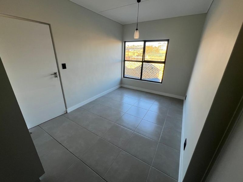 A bright, empty bedroom featuring neutral grey floor tiles, a modern pendant light, and a large window with black framing.
