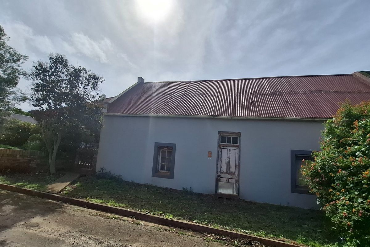 Front elevation of a cottage-style house featuring a corrugated metal roof, timber door, and timber-framed windows, showing signs of exterior weathering.