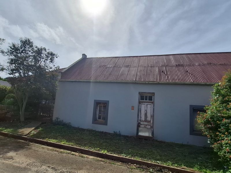 Front elevation of a cottage-style house featuring a corrugated metal roof, timber door, and timber-framed windows, showing signs of exterior weathering.