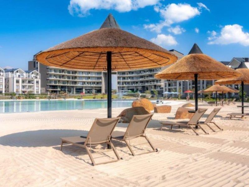 A well-maintained communal pool area featuring a sandy beach entry, thatched umbrellas, and modern apartment buildings in the background.