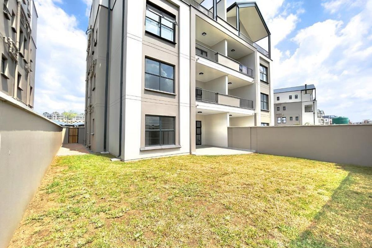 Rear view of a modern apartment building featuring private balconies, a paved patio area, and a private garden space with perimeter walling.