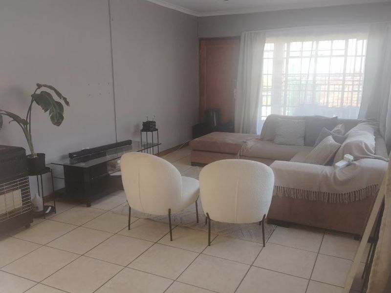 A living room featuring ceramic floor tiles, ceiling track lighting, and a large window with security gate, offering a functional and neutral living space.