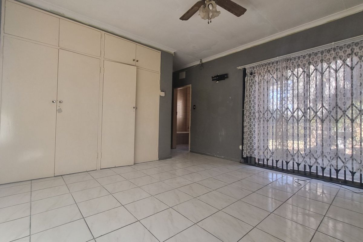 A spacious bedroom featuring tiled flooring, extensive built-in cupboards, a ceiling fan, and a security gate leading to the exterior.