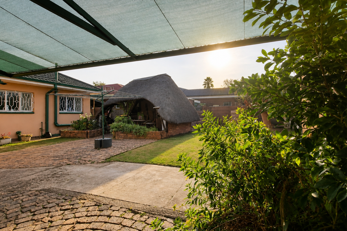 A spacious rear garden area featuring a traditional thatched lapa, paved surfaces, and a large shade cloth canopy providing outdoor cover.