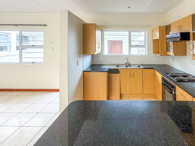 An open-plan kitchen and living area featuring light wood cabinetry, dark granite countertops, built-in appliances, and neutral tiled flooring.