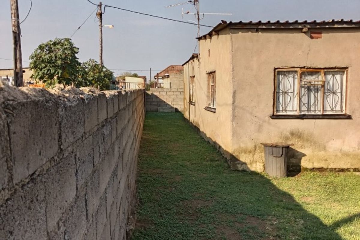 A side view of the property showing a narrow grass yard, concrete boundary wall, and exterior wall with metal-framed windows and security bars.