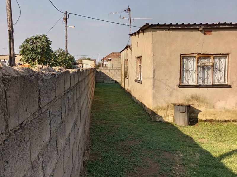 A side view of the property showing a narrow grass yard, concrete boundary wall, and exterior wall with metal-framed windows and security bars.