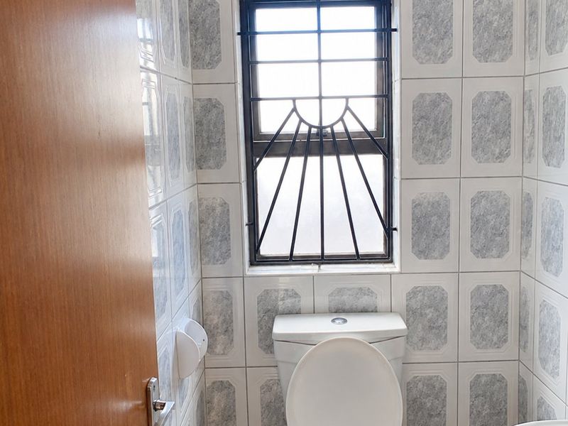 A compact bathroom featuring a standard toilet, washbasin, and full-height wall tiling with a security-barred window for ventilation.