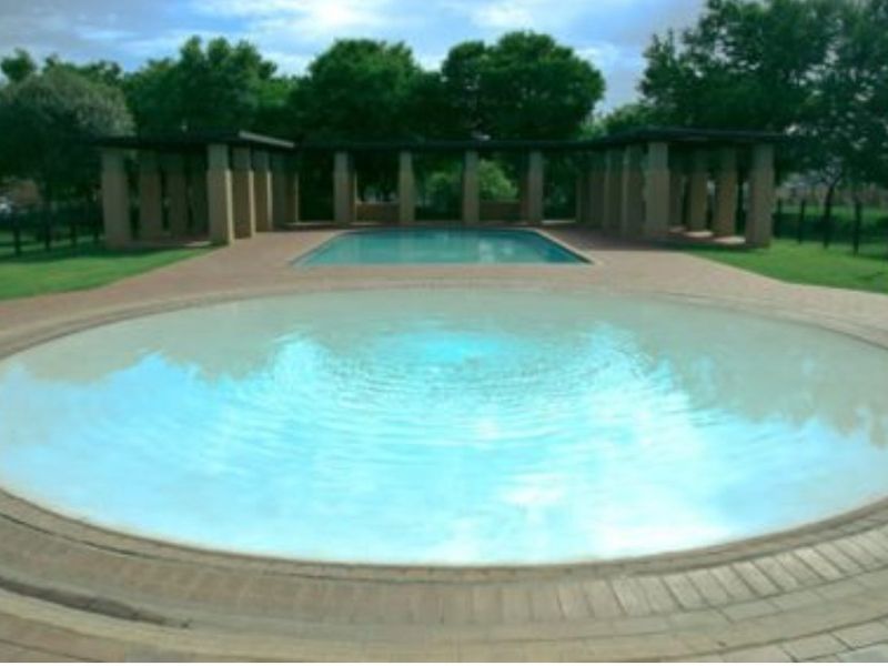 A communal outdoor pool area featuring a circular pool, a secondary rectangular pool, and a covered pergola structure set within a landscaped garden.