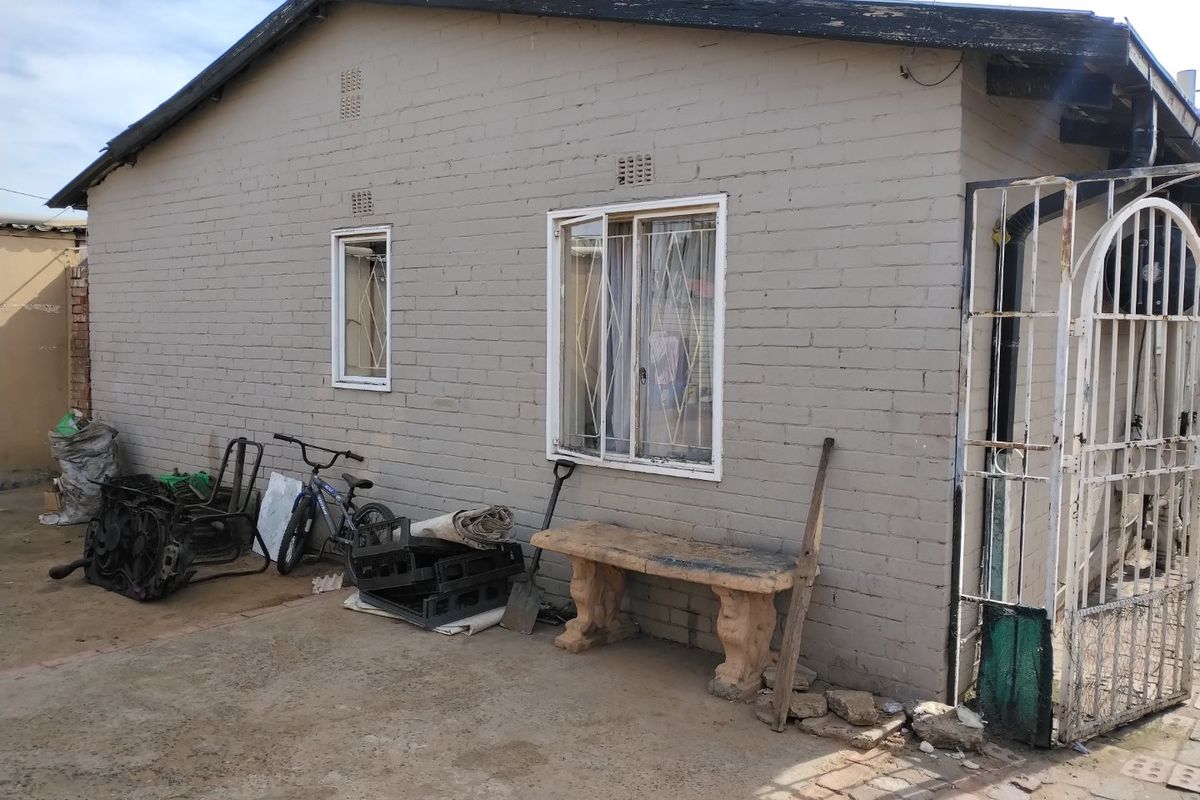 Rear exterior view of a brick house featuring security bars on windows, a metal gate, and concrete paving in need of maintenance.
