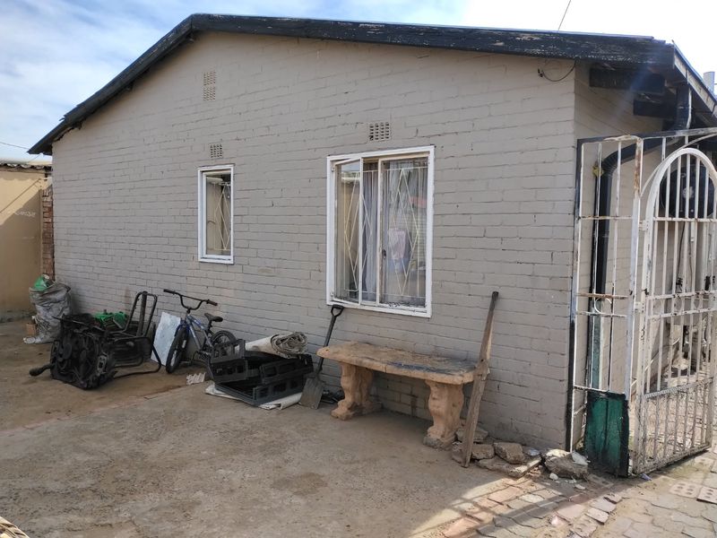 Rear exterior view of a brick house featuring security bars on windows, a metal gate, and concrete paving in need of maintenance.