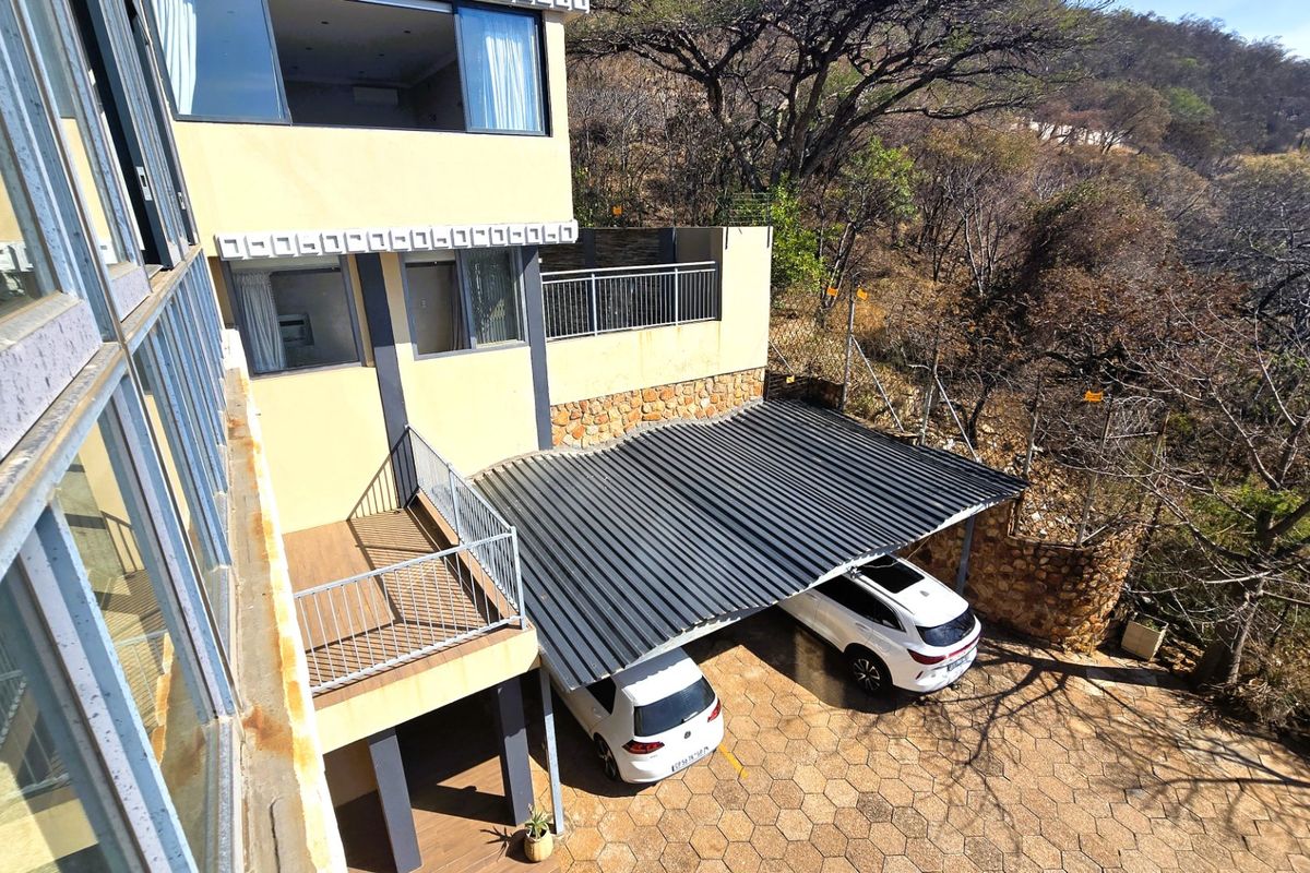 Rear exterior view showing a multi-level house facade, a corrugated metal carport, stone retaining walls, and a paved driveway area.