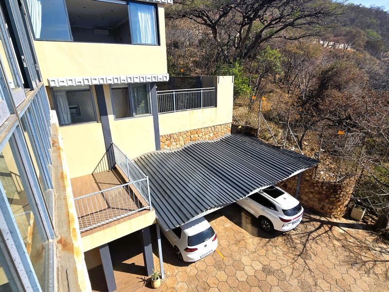 Rear exterior view showing a multi-level house facade, a corrugated metal carport, stone retaining walls, and a paved driveway area.