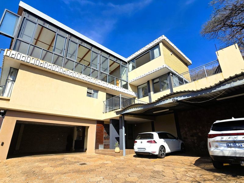 Front elevation of a multi-level house featuring a paved driveway, double garage, carport, and an upper-level balcony with extensive glass windows.