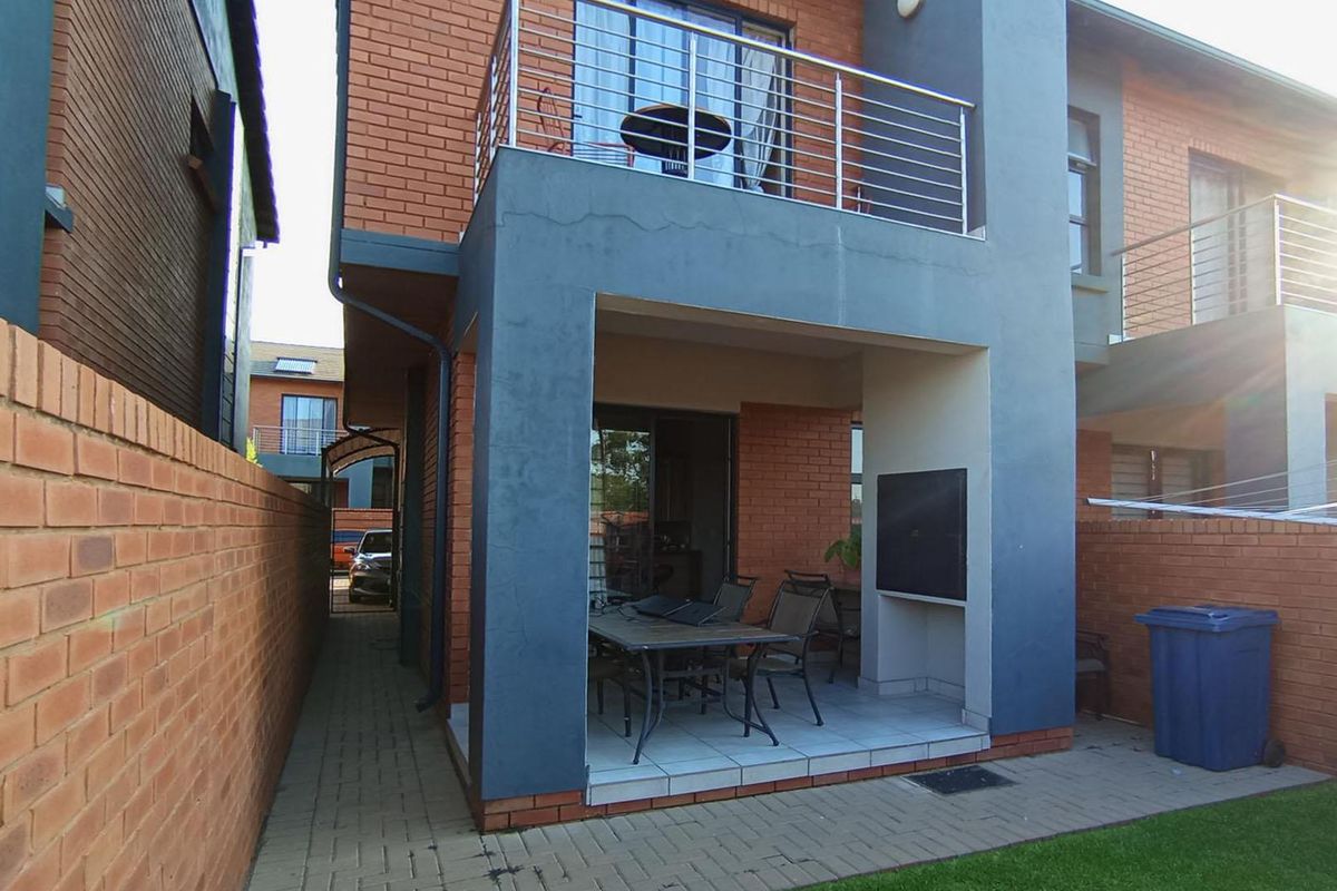 Rear view of the townhouse featuring a covered patio with a built-in braai, a paved garden area, and an upper-level balcony with metal railings.