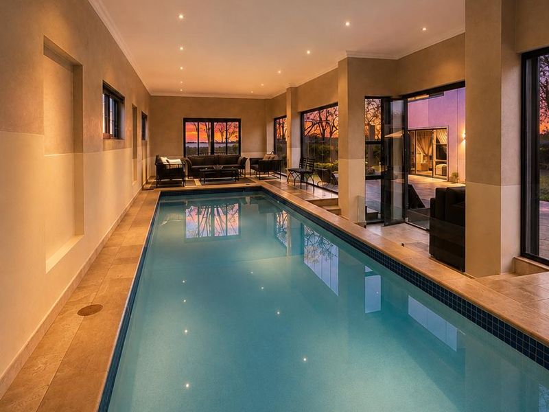 A luxurious indoor lap pool area featuring tiled surrounds, recessed lighting, and large glass doors opening to an outdoor patio with sunset views.