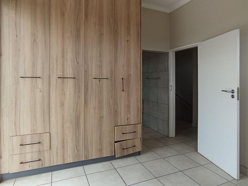 A bright, empty bedroom featuring modern wood-finish built-in cupboards, tiled flooring, and direct access to an adjoining space.