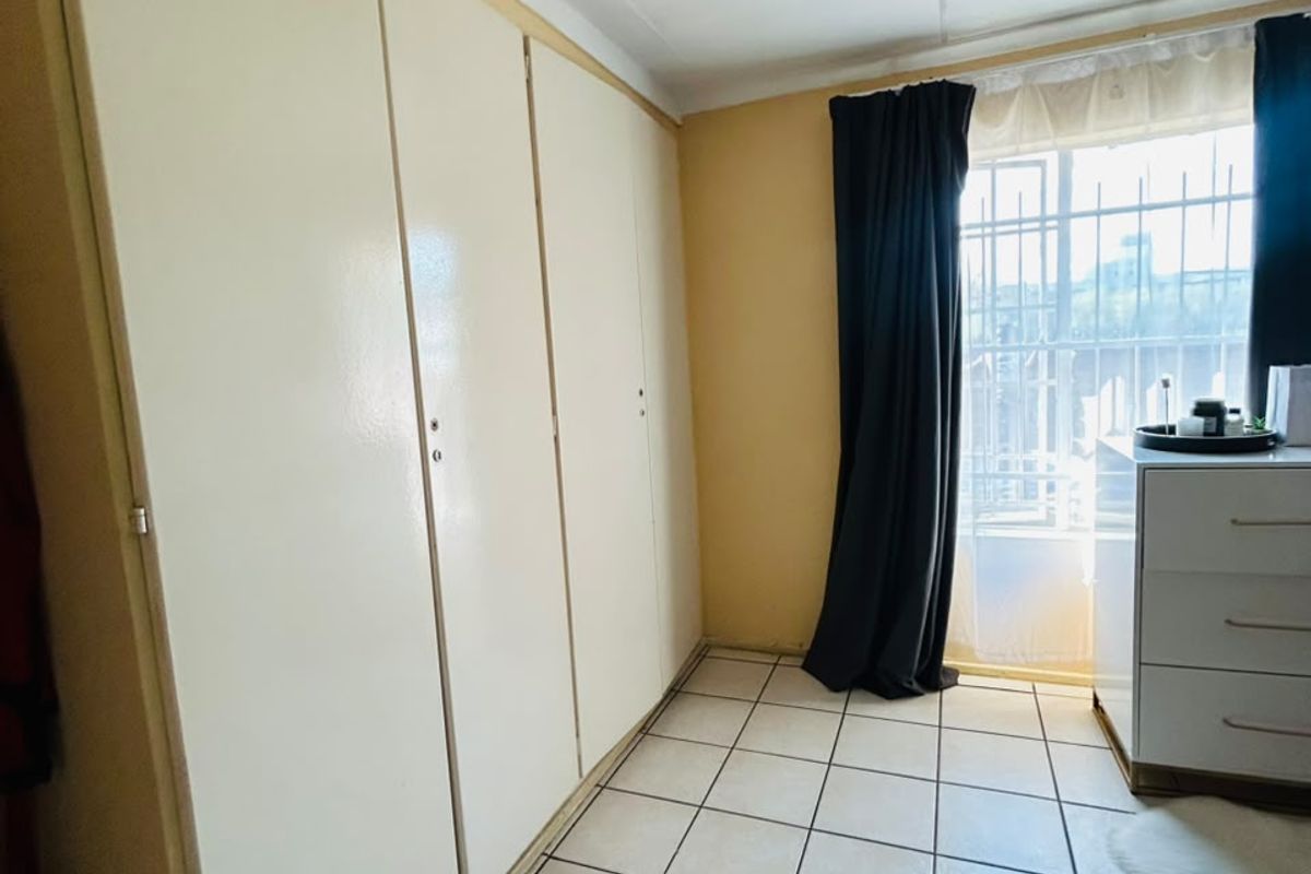 A bedroom featuring built-in cupboards, tiled flooring, and a window fitted with security bars, offering a functional and secure living space.