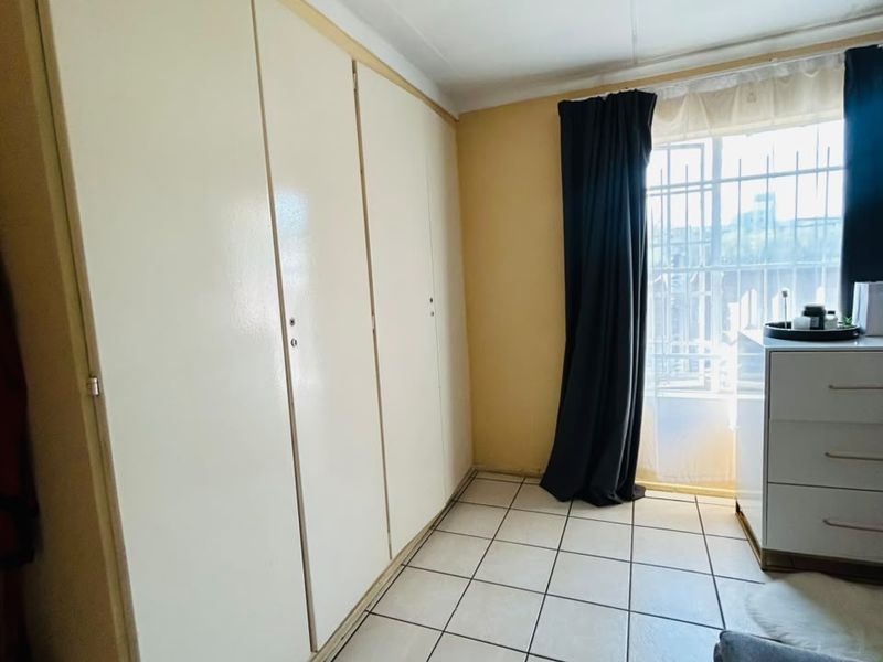 A bedroom featuring built-in cupboards, tiled flooring, and a window fitted with security bars, offering a functional and secure living space.