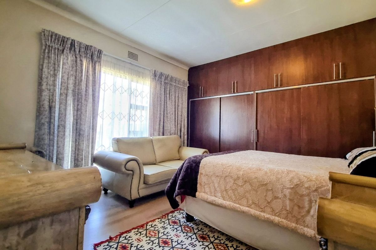 A spacious bedroom featuring extensive built-in wooden cupboards, laminate flooring, and large windows with curtain rails.